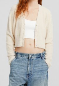 White cropped cardigan made of soft, fluffy material with six buttons down the front, paired with blue high-waisted jeans.