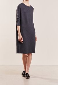 Donna che indossa un vestito a maglia grigio scuro fino al ginocchio con maniche a tre quarti e mocassini neri in pelle lucida con dettagli in oro.