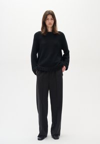 Črni pleteni pulover z teksturirano zasnovo, sproščenega kroja in rebrastimi manšetami. V kombinaciji z elegantnimi črnimi hlačami in črnimi čevlji s koničastim nosom.