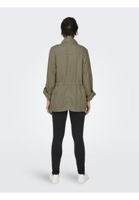 Veste vert olive avec taille élastique, manches retroussées et coupe décontractée, associée à des leggings noirs et des baskets blanches.
