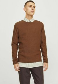 Suéter marrón texturizado de manga larga, con un cuello redondo, combinado con una camisa de cuadros de botones, junto con pantalones oscuros.