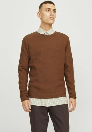 Pull en maille marron avec des manches longues, un col rond, superposé sur une chemise à carreaux avec boutons, associé à un pantalon sombre.