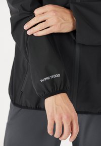 Svart vattentät jacka med elastiska muddar, försedda med "W-PRO 15000" tryckt i vitt på vänster ärm. Slät, matt tygtextur.