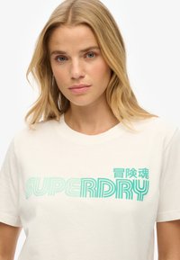 Kremowa bawełniana koszulka z grafika w kolorze teal z napisem "SUPERDRY", falistymi liniami i dodatkowymi znakami po lewej stronie.
