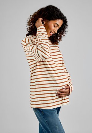 Terhes nő csíkos hosszú ujjú pólóban és farmerben, gyengéden tartja a hasát, mosolyog és lefelé néz egyszínű háttér előtt.