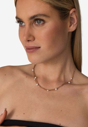 Junge Frau mit heller Haut und blonden Haaren trägt eine doppelte Goldkette mit kleinen weißen Perlen und schaut nach rechts.