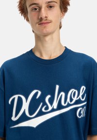 Blå bomullströja med en stor vit grafik av texten "DC shoe Co.", med korta ärmar och rundad halsringning. Mjuk textur.