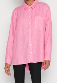 Pink linen shirt with a button-down collar, long sleeves, single chest pocket, and a slightly longer back hem. Textured fabric finish.