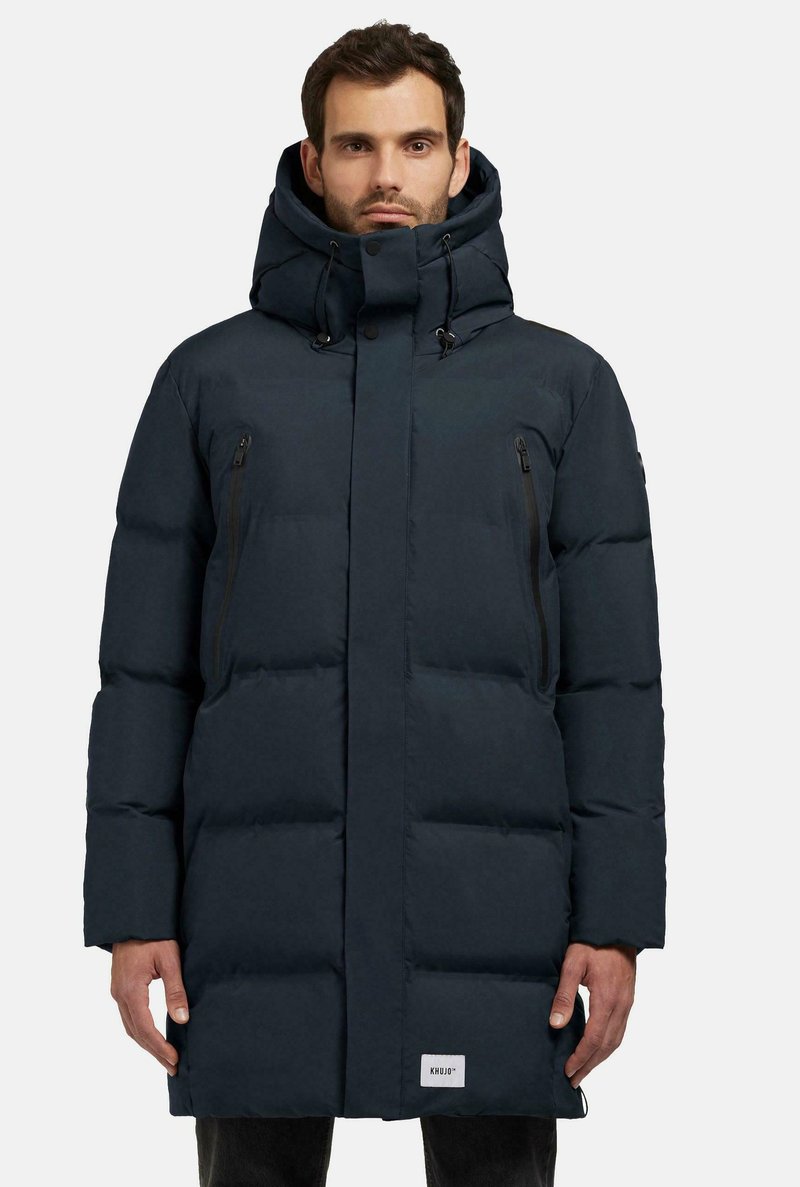 Mørkeblå polstret jakke med høj krave og justerbar snoretræk-hætte. Har to lynlåslommer på brystet og et logo-patch på kanten.