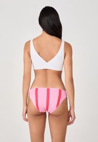 Donna in piedi con la schiena rivolta verso la fotocamera, indossante un reggiseno bianco e delle mutande rosa a righe verticali rosse, su uno sfondo neutro.