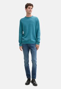 Türkiser Pullover mit langen Ärmeln und rundem Ausschnitt, getragen über blauen Jeans und schwarzen Schuhen. Stricktextur mit lockerem Schnitt.