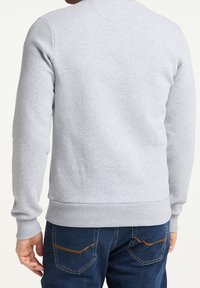 Sudadera gris de cuello redondo con puños y dobladillo acanalados, hecha de una tela suave. Vista desde la parte de atrás, con jeans azul oscuro y costuras de acento naranjas.