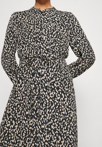 Donna che indossa un vestito nero a maniche lunghe con macchie astratte beige e bianche, con bottoni sul davanti e cintura legata in vita, mani dietro la schiena.