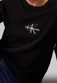 Černý svetřík s kulatým výstřihem vyrobený z texturované látky, s bílým logem Calvin Klein Jeans a velkým designem "CK" na přední straně.