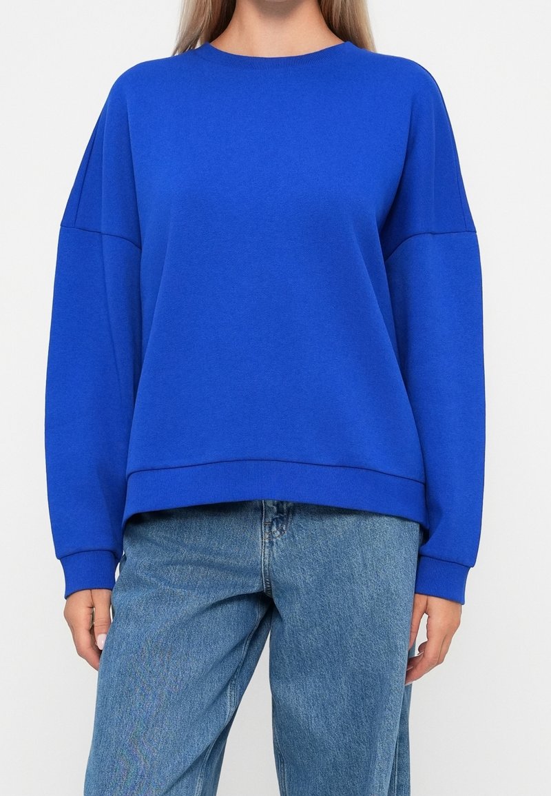 Femme portant un sweat bleu royal ample et un jean bleu clair, montrée des épaules jusqu’aux hanches sur fond blanc.