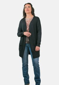 Larga chaqueta negra hecha de material suave, con un corte relajado, abertura frontal y dos bolsillos delanteros, llevada sobre una blusa con estampado.