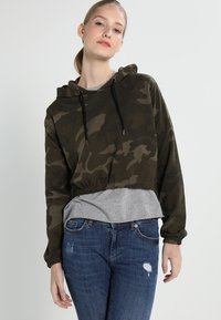 Camo-patterned cropped hoodie in dark green with a drawstring hood, elastic hem, long sleeves, layered over a gray shirt, paired with blue jeans.