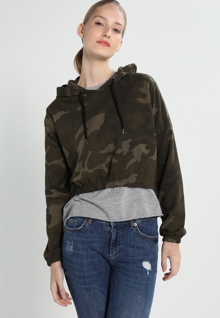 Camo-patterned cropped hoodie in dark green with a drawstring hood, elastic hem, long sleeves, layered over a gray shirt, paired with blue jeans.