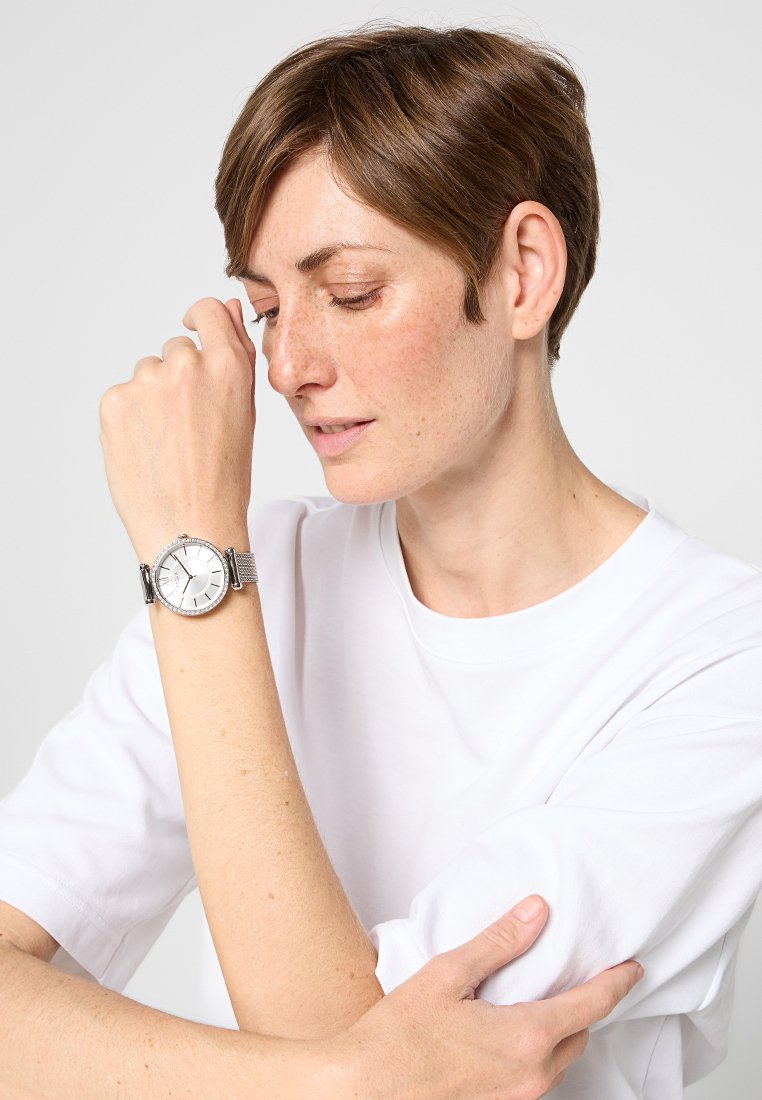 Reloj de metal plateado con una esfera blanca redonda. Presenta un bisel texturizado y una pulsera de eslabones. Se lleva en un brazo desnudo con una camisa de manga corta blanca.