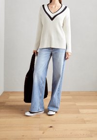 Person med vit ribbad V-ringad tröja med mörka ränder, ljusblå vida jeans, svart-vita sneakers, som håller i en svart stickad väska.