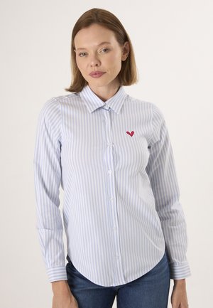 Femme portant une chemise à rayures bleu clair et blanc à boutons avec un petit cœur rouge brodé sur la poitrine, associée à un jean bleu.