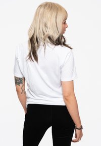 White cotton t-shirt with short sleeves, relaxed fit, showing smooth texture; worn with fitted black pants. Visible tattoo on left arm.