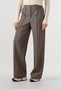 Kobieta ubrana w brązowe spodnie o szerokich nogawkach oraz beżową teksturowaną bluzkę z długim rękawem, w parze z czarnymi, białymi i metalicznymi sneakersami, stojąca na białej podłodze.