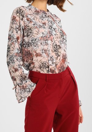 Blouse florale rose clair avec des fleurs noires et orange, dotée d'un col froncé et de poignets volantés, associée à un pantalon rouge taille haute.