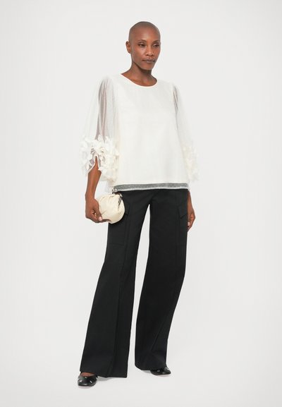 Bela majica s prosojnimi rokavi, okrašenimi z ro�žami, v kombinaciji s črnimi širokimi hlačami. Manekenka drži majhno kremo torbico.
