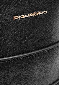 Bolso de cuero negro con superficie texturizada, la marca "Piquadro" en dorado y detalles de costuras lisas en los bordes.