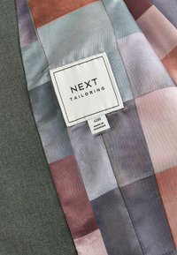 Interior de chaqueta a medida con un forro a rayas multicolor en tonos de azul, rosa y beige; presenta una etiqueta blanca que dice "NEXT TAILORING".