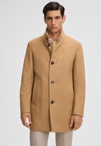 Cappotto di lana beige con colletto alto, cinque bottoni, due tasche anteriori e taglio dritto. Il tessuto ha una consistenza liscia e una vestibilità sartoriale.