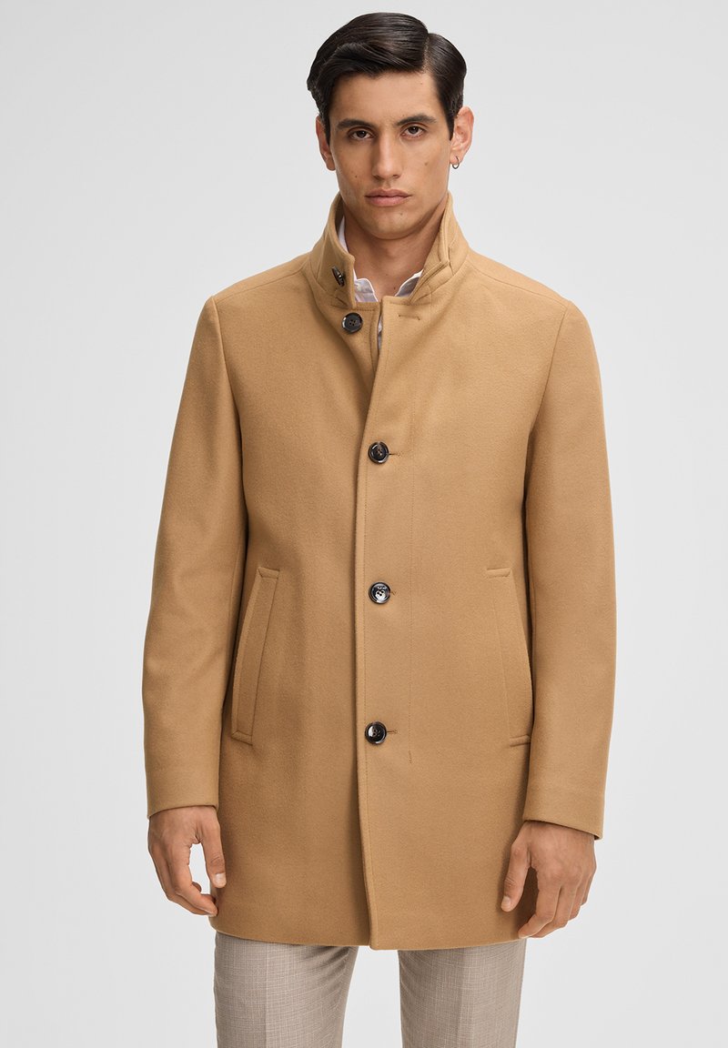 Cappotto di lana beige con colletto alto, cinque bottoni, due tasche anteriori e taglio dritto. Il tessuto ha una consistenza liscia e una vestibilità sartoriale.