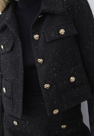 Veste noire texturée avec de gros boutons dorés en forme de fleurs et jupe assortie, portées sur un pull à col roulé côtelé noir.