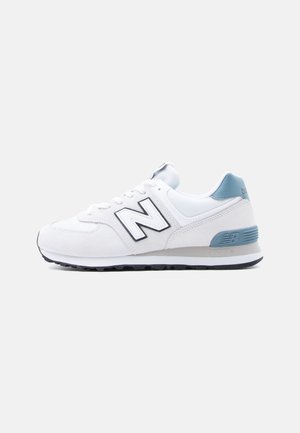 Hvit og lyseblå New Balance-sneaker med svart logo, skolisser og en teksturert svart såle vist fra siden på en hvit bakgrunn.