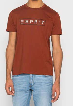 Homme portant un T-shirt à manches courtes couleur rouille avec le logo "ESPRIT" et un jean bleu clair, debout devant un fond uni.