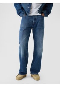 Jean en denim bleu avec une coupe décontractée, coutures visibles et légère décoloration. Assorti à des chaussures de style mocassin de couleur beige.