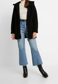 Cappotto in fleece nero con cappuccio, chiusura a zip e tasche laterali, indossato sopra un topcremoso a coste e jeans blu svasati con orli grezzi. Stivaletti neri alla caviglia.