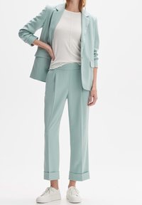 Un costume de mint clair composé d'un blazer ajusté avec des manches retroussées et un pantalon assorti, complété par un haut crème et des baskets blanches.