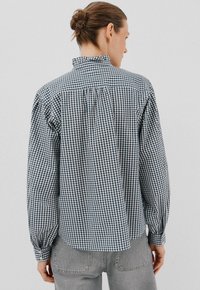 Vrouw met bruin haar in een knot, gekleed in een zwart-wit geruite blouse met lange mouwen en grijze spijkerbroek, met haar gezicht van de camera weg tegen een effen achtergrond.