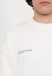 Hvit hettegenser laget av økologisk bomull, med teksturert rund hals og trykte vaskeanvisninger på venstre side.