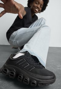 Une personne souriante portant un jean bleu clair et des baskets noires, assise par terre, tendant la main vers l'appareil photo.