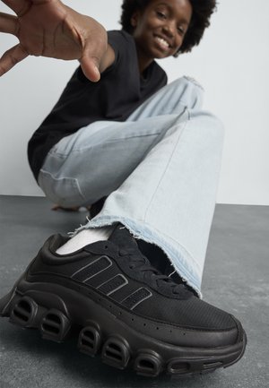 Une personne souriante portant un jean bleu clair et des baskets noires, assise par terre, tendant la main vers l'appareil photo.