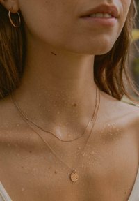 Rosa goldene, geschichtete Halsketten mit einem strukturierten Anhänger, die auf Haut mit Wassertropfen präsentiert werden und den Glanz sowie die Details des Schmucks hervorheben.