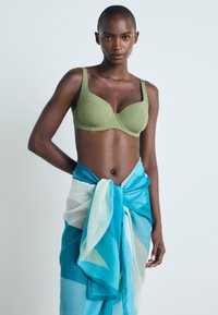 Sostén verde con copas estructuradas, combinado con una falda cruzada de patrón azul y blanco atada a la cintura, sobre un fondo neutro.