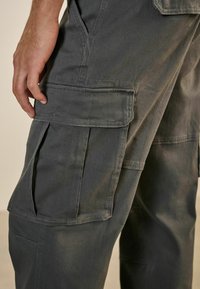 Pantalones cargo grises con múltiples bolsillos laterales, fabricados en un tejido duradero. La textura es suave con costuras visibles y un diseño de ajuste relajado.