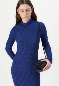 Rochie tricotată cu mâneci lungi, de un albastru profund, cu un model geometric texturat; modelul ține o mică geantă de mână neagră, cu o formă curbată și o curea.