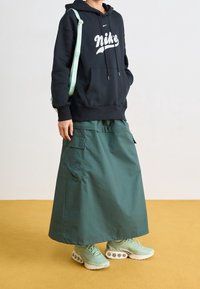 Person klädd i svart Nike-hoodie, lång grön cargokjol, mintgröna sneakers och bärande på en ljusgrön axelväska på en gul golv.