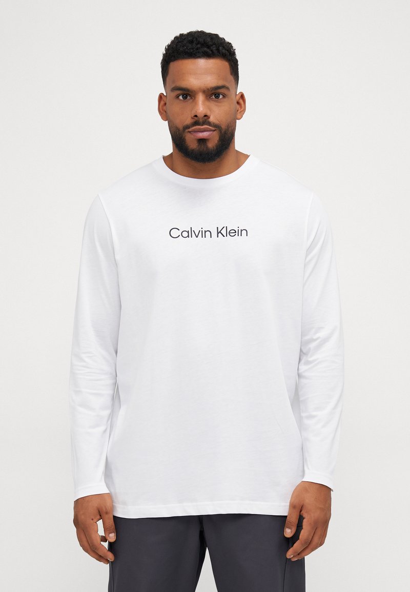 Fehér, hosszú ujjú póló, fekete "Calvin Klein" logóval, puha anyagból készült, laza szabású és kerek nyakú.
