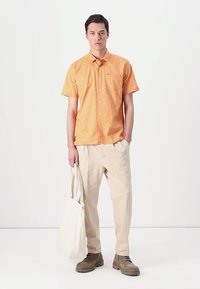 Camisa laranja de manga curta com fecho de botão à frente e bolso no peito, combinada com calças bege e uma bolsa de ombro branca. Sapatos em tons terrestres.